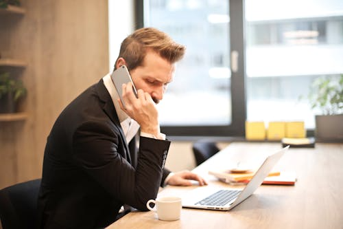 A man in a suit talking on a phone while working on his laptop in his office