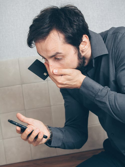A man covering his mouth in shock with a phone in one hand and a credit card in the other