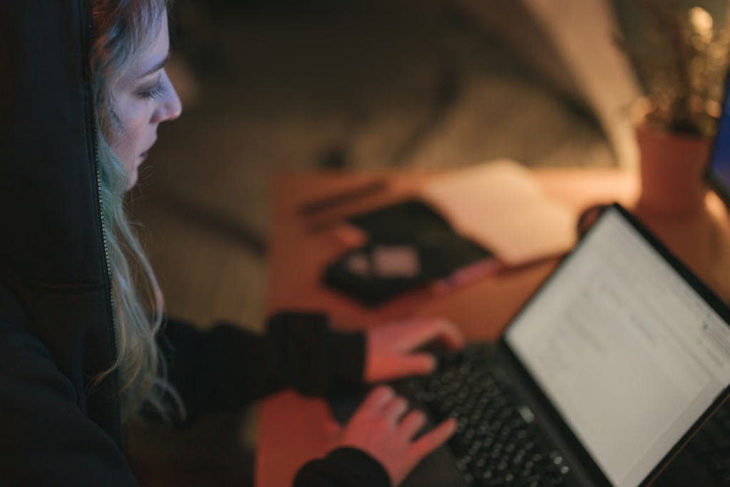 A person in a dark hoodie typing on a laptop in a dimly lit setting