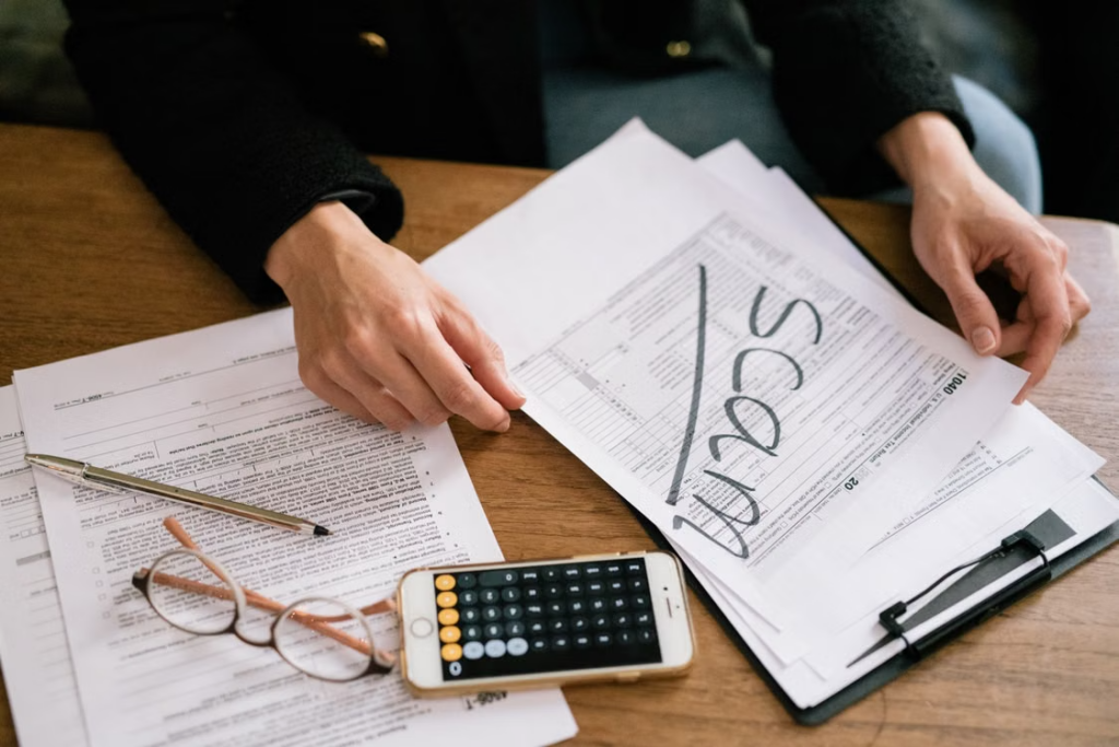 A person reviewing tax documents with the word SCAM written across the top sheet next to a calculator and glasses
