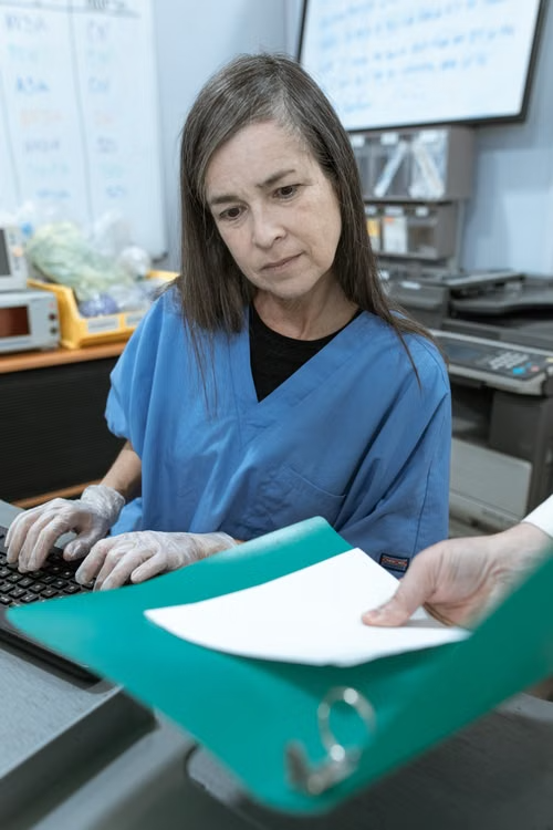 A nurse in blue scrubs typing at a keyboard while reviewing documents in a binder