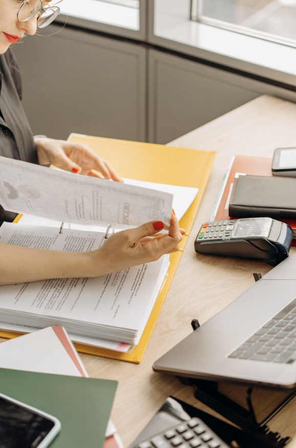 This photo shows a woman reviewing printed documents beside a laptop.