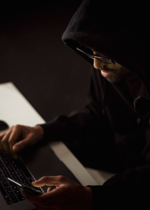 This image shows a hooded hacker using a laptop and smartphone.